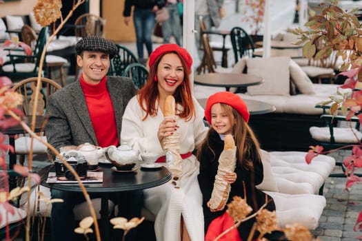 A stylish family of three is sitting at a table outside in a cafe and drinking coffee. Dad, mom and daughter in the autumn city.