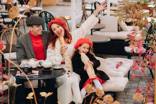 A stylish family of three is sitting at a table outside in a cafe and drinking coffee. Dad, mom and daughter in the autumn city.