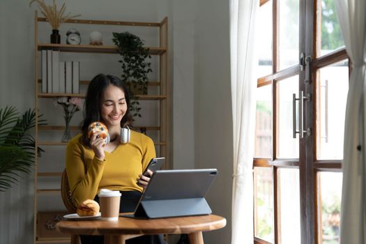Beautiful Asian woman is using laptop, drinking coffee and smiling while sitting by the window at home...
