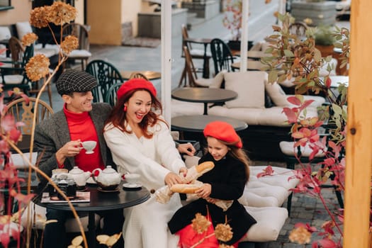 A stylish family of three is sitting at a table outside in a cafe and drinking coffee. Dad, mom and daughter in the autumn city.