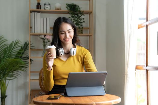 Beautiful Asian woman is using laptop, drinking coffee and smiling while sitting by the window at home...