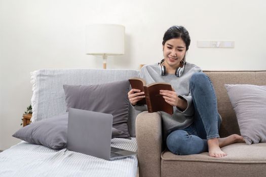 Young beautiful asian woman student sitting on sofa reading book at home while using laptop for online learning