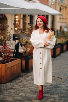 Portrait of a pretty woman in a red beret and a white cardigan with baguettes in her hands strolling through the autumn city.