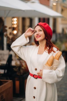Portrait of a pretty woman in a red beret and a white cardigan with baguettes in her hands strolling through the autumn city.