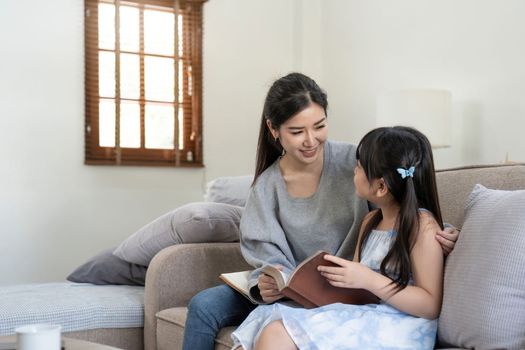 happy asian woman reading a book to daughter embracing her at home. sitting on sofa.
