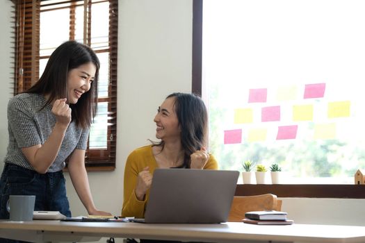 Two young Asian businesswomen show joyful expression of success at work smiling happily with a laptop computer in a modern office..