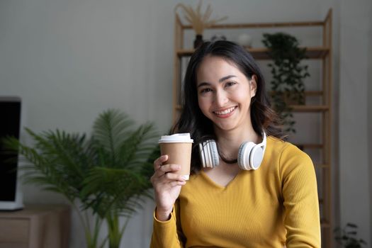 Beautiful Asian woman is using laptop, drinking coffee and smiling while sitting by the window at home...