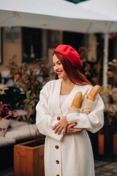 Portrait of a pretty woman in a red beret and a white cardigan with baguettes in her hands strolling through the autumn city.