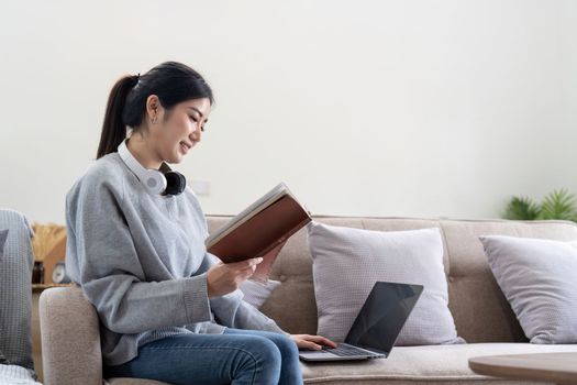 Young beautiful asian woman student sitting on sofa reading book at home while using laptop for online learning.