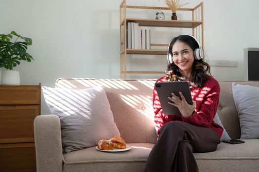 Beautiful woman asian wearing sweater listening music on line with a tablet sitting on a sofa in the living room at home