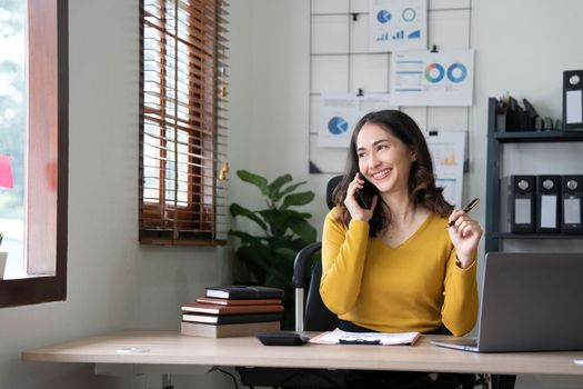 Smiling beautiful Asian businesswoman analyzing chart and graph showing changes on the market and holding smartphone at office..