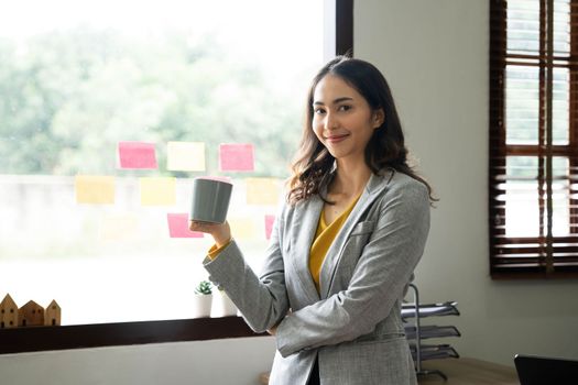 Beautiful young Asian businesswoman smiling holding a coffee mug and laptop working at the office..