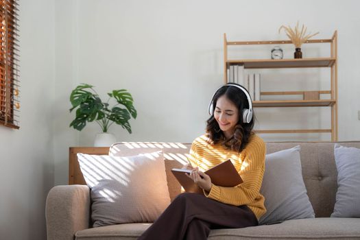 Young Asian woman listening music from headphones and writing note for her work idea in diary book.She sitting on grey sofa in living room..