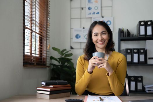 Beautiful young Asian businesswoman smiling holding a coffee mug and laptop working at the office..