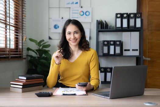 Smiling beautiful Asian businesswoman analyzing chart and graph showing changes on the market and holding smartphone at office..