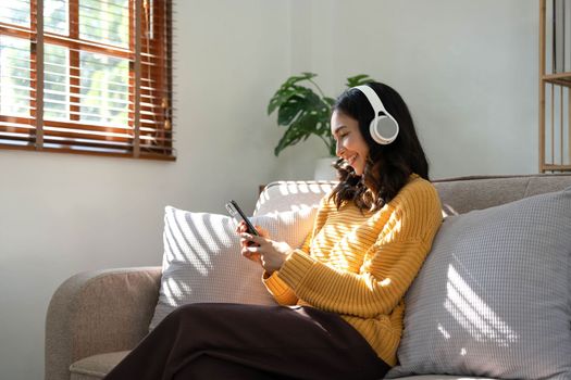 Smiling young woman sit on sofa at home talk on video call on smartphone with headphones. Happy millennial female in earphones use cellphone have webcam digital online communication..