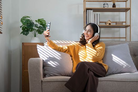 Smiling young woman sit on sofa at home talk on video call on smartphone with headphones. Happy millennial female in earphones use cellphone have webcam digital online communication..