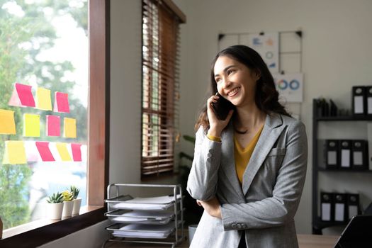 Smiling beautiful Asian businesswoman analyzing chart and graph showing changes on the market and holding smartphone at office..