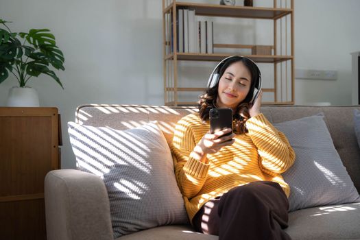 Young Asian woman listening music from headphones and writing note for her work idea. She sitting on grey sofa in living room..