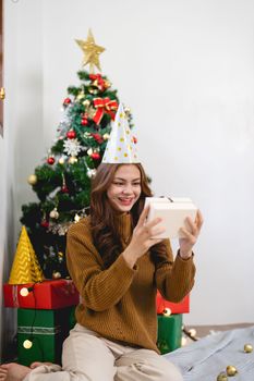 Exciting woman sitting against Christmas tree background. Cheerful lady surprised of the present after the opening in the gift box. Marry Christmas and Happy Holidays.