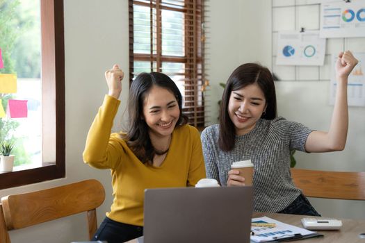 Two young Asian businesswomen show joyful expression of success at work smiling happily with a laptop computer in a modern office..