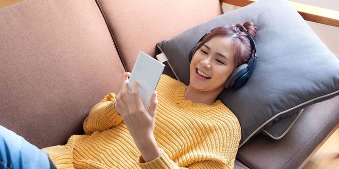 Young beautiful Asian woman laying in sofa and playing smartphone with headphones in home background.Concept of people using technology.Vintage tone.