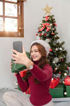 Holiday selfie at Christmas tree. Happy young girl taking selfie on smart phone at decorated Christmas tree iat home.
