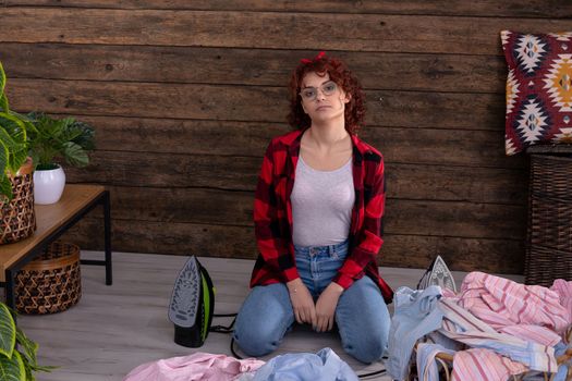 A young girl is waiting to iron men's shirts. She sits on the floor of a room in the house. An overturned basket of clothes. A wall of brown boards.