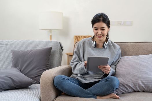 Happy pretty young woman in casual outfit sitting on sofa at home and using tablet while browsing internet.