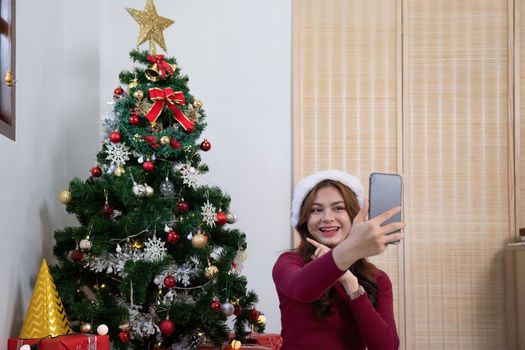 Young woman using smart phone video call talking. Christmas tree decorated with ornament in living room at home. Christmas and New Year holiday festival