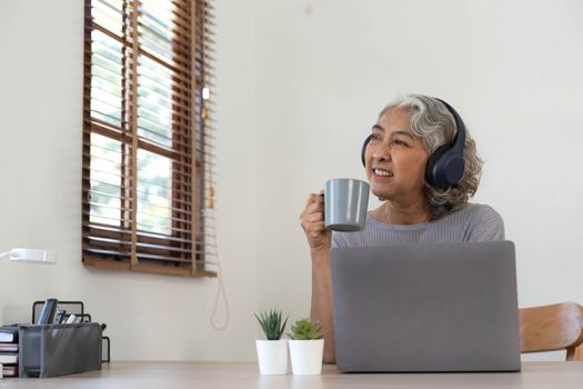 Senior woman using laptop while wearing headphones at home - Joyful elderly lifestyle and technology concept.