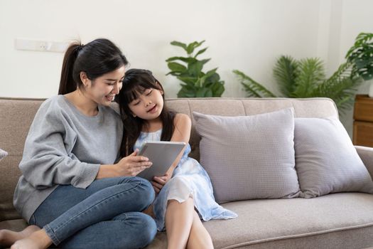 happy asian woman showing digital tablet to daughter embracing her at home. sitting on sofa.