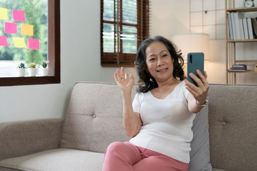 Asian elderly woman taking selfie on mobile phone while sitting on sofa and smiling.