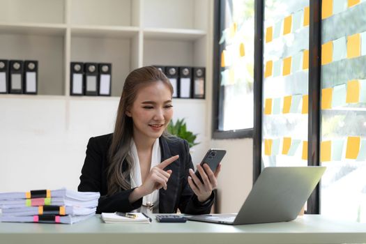 Smiling beautiful Asian businesswoman analyzing chart and graph showing changes on the market and holding smartphone at office...