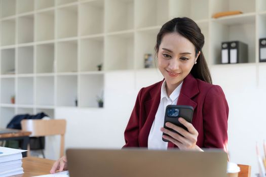 Smiling beautiful Asian businesswoman analyzing chart and graph showing changes on the market and holding smartphone at office..