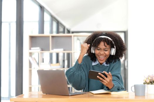 Photo of cheerful joyful mixed race woman playing games and wearing wireless headphones at home Play game on your smartphone that wins the moment of victory. Fun and enjoyable vacation.