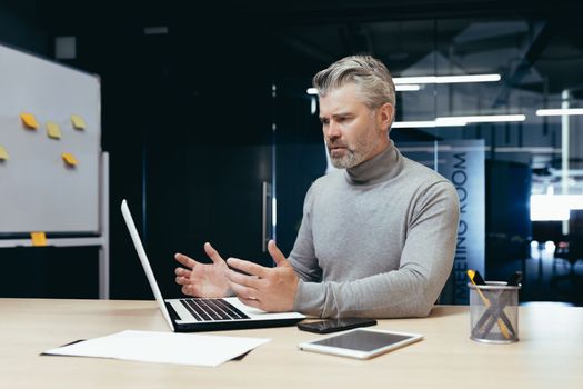 Dissatisfied gray haired boss in office, senior man disappointed with achievement result, mature businessman upset and sad working with documents and using laptop inside building.