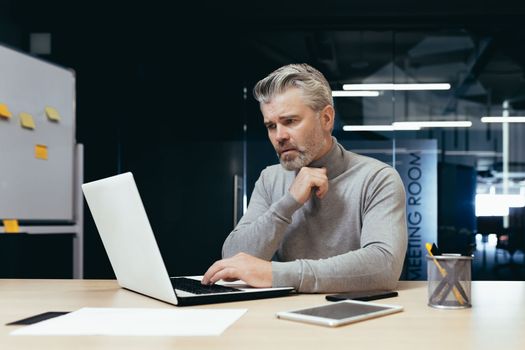 Serious thinking businessman inside office at work with laptop, senior gray haired man working sitting at desk.