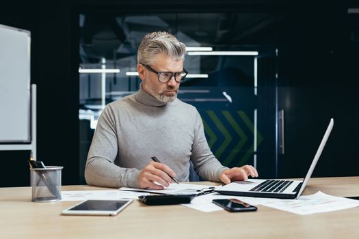 Serious thinking businessman inside office at work with laptop, senior gray haired man working sitting at desk.