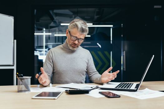 Dissatisfied gray haired boss in office, senior man disappointed with achievement result, mature businessman upset and sad working with documents and using laptop inside building.