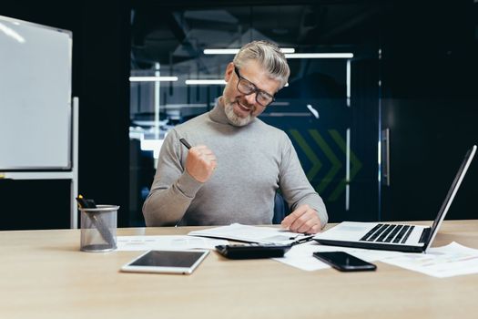 Happy senior business man sitting in the office at the table and signing a successful contract. making a profitable deal. Happy shows with hand yes.