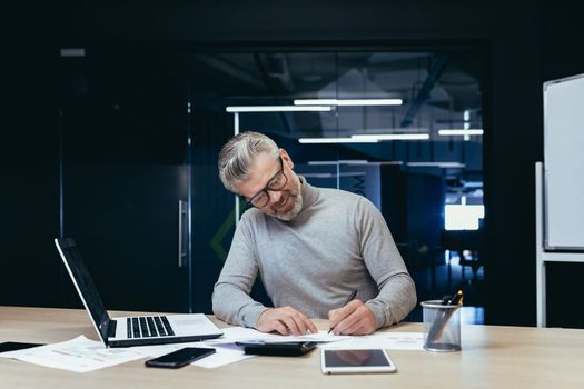 Senior mature financier investor working smiling and satisfied with documents, mature businessman on paper work with invoices and contracts using laptop inside office.