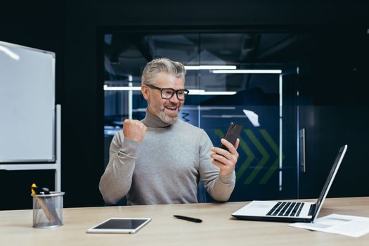 Businessman celebrating victory and success, mature gray haired man inside office got good news result using smartphone, man at work with laptop.