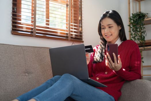 Beautiful young woman holding credit card while using laptop computer and mobile phone sitting at the living room. Black friday online shopping concept.