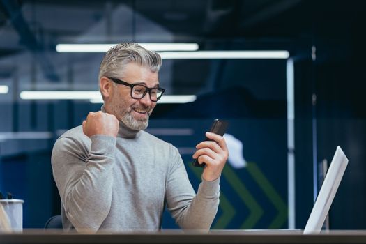 Businessman celebrating victory and success, mature gray haired man inside office got good news result using smartphone, man at work with laptop.