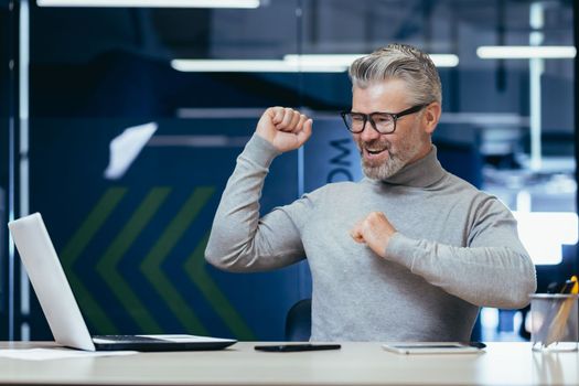 Successful gray haired boss working inside modern office, mature businessman celebrating victory looking at monitor screen and satisfied with achievement, senior man smiling and dancing sitting.