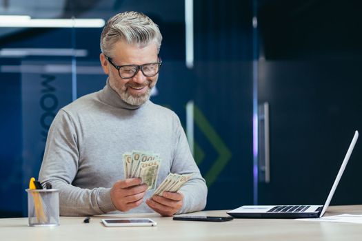 Mature successful financier investor counts money dollars cash sitting inside office at table, senior businessman happy with result achievement and income uses laptop at work.