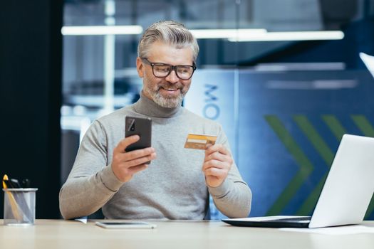 Cheerful and smiling gray-haired businessman inside a modern office makes purchases in an online store, the man holds a bank credit card and a smartphone in his hands, the buyer is satisfied choice.