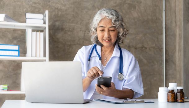 doctor senior grey-haired woman in white medical coat is using a smartphone and smiling while sit use laptop at office.