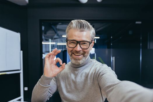 Close-up photo. Senior gray-haired businessman, freelancer takes a selfie, talks to the camera on a video call, shoots a blog. Smiles at the camera, shows ok